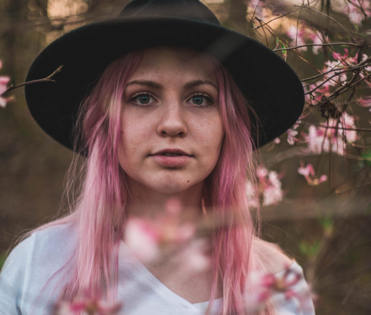 Chica de cabello lacio, largo, despeinado, rosa chicle, con sombrero boho, frente a un árbol de flores rosas