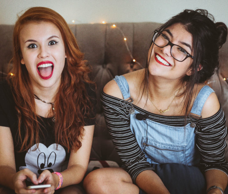 Amigas sentadas en un sillón sonriendo; chica de cabello rojo, largo y ondulado, haciendo gestos, mujer de cabello castaño peinado con chongo, messy bun, con overol