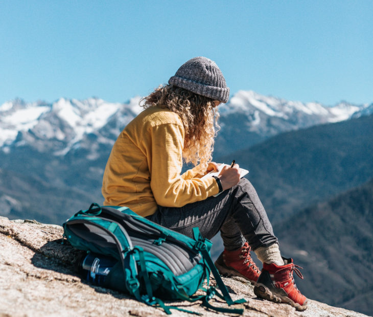 Chica sentada en la cima de una montaña, escribiendo y dibujando en una libreta 