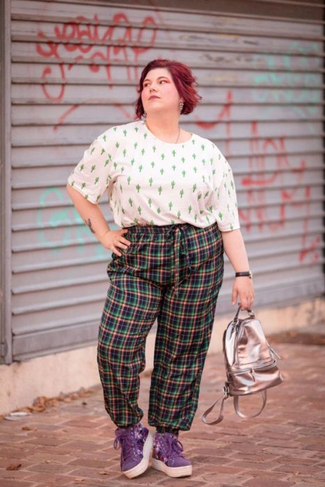 Chica curvy se pone la mano en la cintura y posa con blusa blanca con estampado de cactus y pantalón verde de cuadros