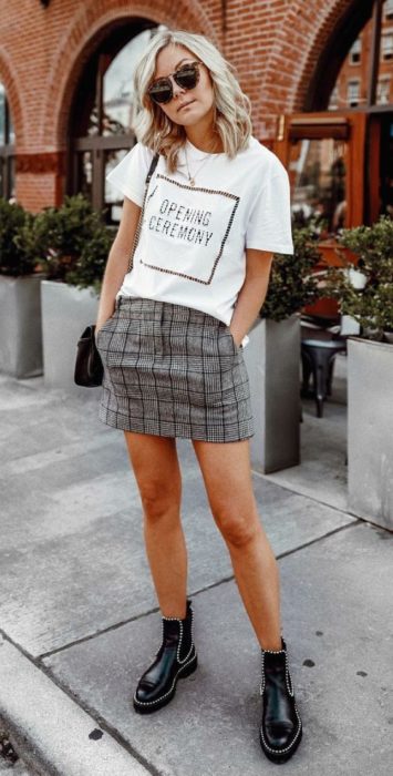 Chica rubia de cabello suelto pone las manos dentro de los bolsillo de su falsa color gris con cuadros y usa una blusa blanca y botines negros