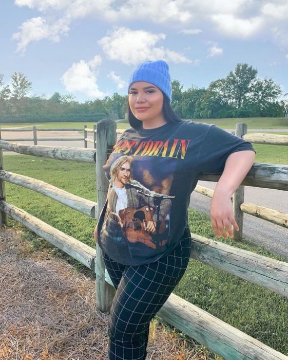 Chica se recarga sobre barda con un gorro azul, blusa amplia negra y pantalón a cuadros color verde