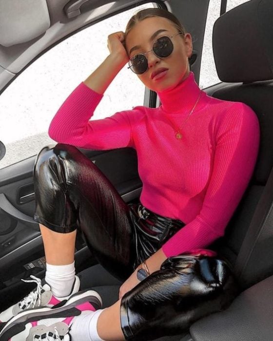Chica posando dentro de una automóvil con pantalón de latéx negro y blusa de cuello alto rosa neón