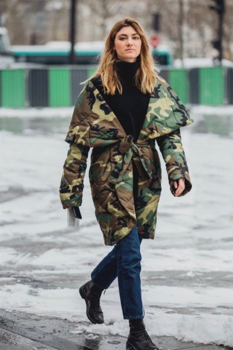 Chica caminando por la calle mientras usa jeans, blusa negra, botas y una chaqueta verde militar 