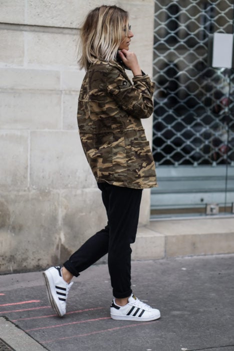 Chica caminando por la calle mientras usa jeans, blusa negra, botas y una chaqueta verde militar 
