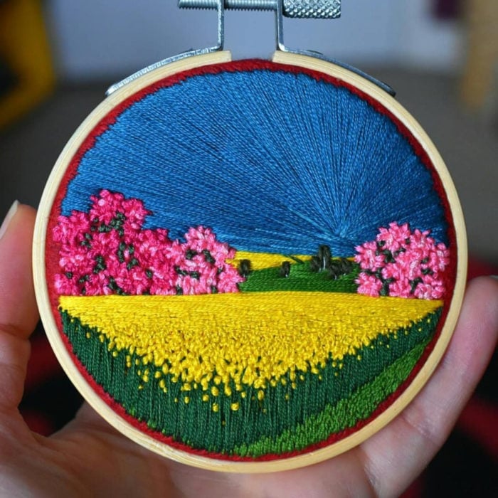 Artista Victoria Rose Richards hace bordados de paisajes naturales; campo de flores amarillas, árboles rosas y cielo azul