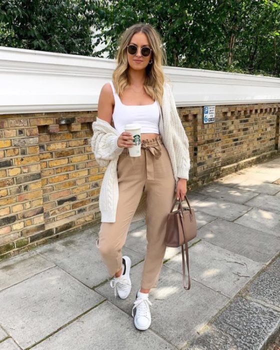 Chica rubia con lentes de sol con top blanco y paper bag pants beige con una café en la mano