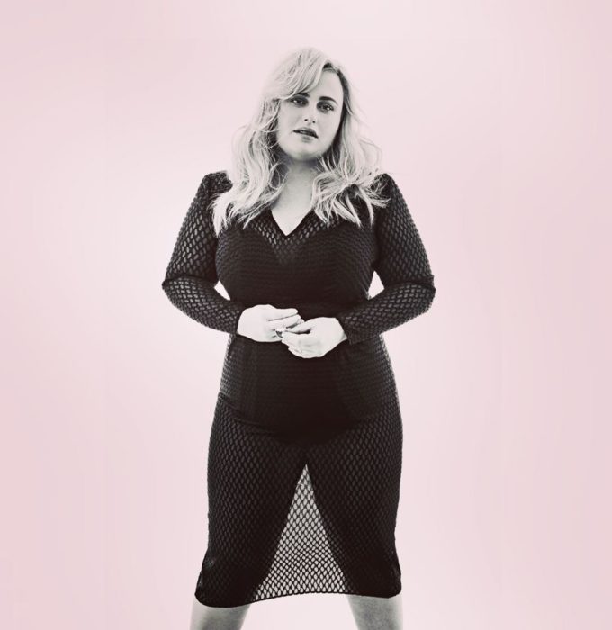 Rebel Wilson posando con ropa negra deportiva y un blusón estilo maya sobre un fondo roja pastel 