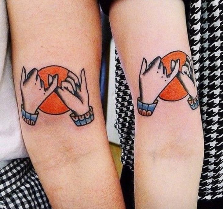 Tatuajes para compartir con tus amigas, antebrazos con diseño de manos entrelazando lso pulgares frente a un sol naranja
