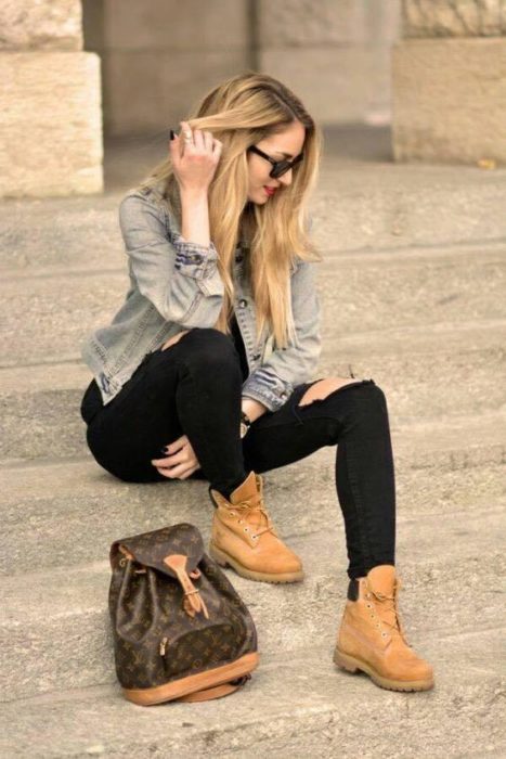 Mujer rubia sentada en escalones se toca el cabello con la mano derecha y viste pantalón negro, chamarra de mezclilla y botas tipo tomberland color camel