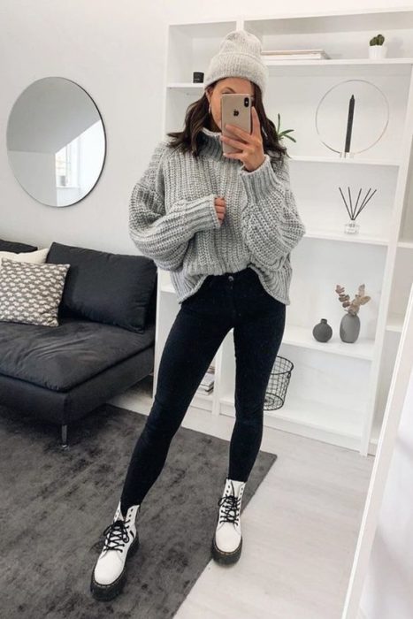 Chica de melena media se toma selfie frente al espejo con suéter gris oversized, pantalón negro y botas dr. martens blancas
