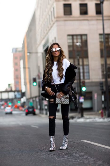 Chica de cabello largo castaño ondulado parada en la calle con pantalón y chamarra negros, blusa blanca y botines de animal print