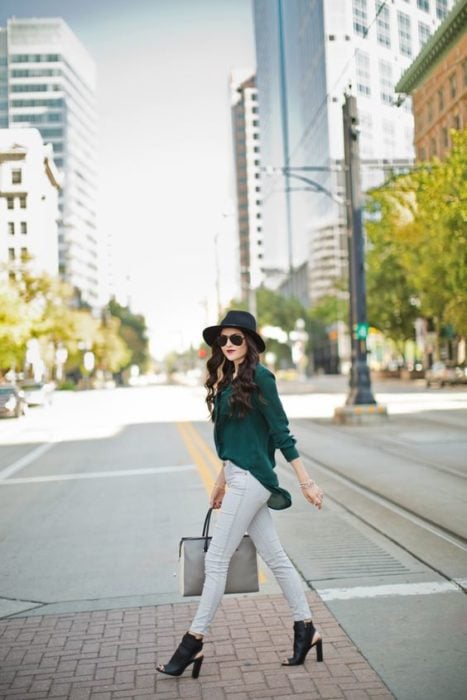 Chica elegante atraviesa la cale con pantalones de mezclilla claros, blusa verde esmeralda, sombrero negro y botines con abertura en la parte de adelante