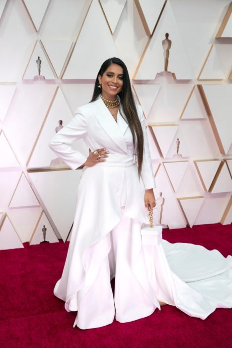 Lily Singh usando un pantalón y una chaqueta de color blanco con una cauda larga y un bolso cuadrado del mismo color durante los Óscar 2020