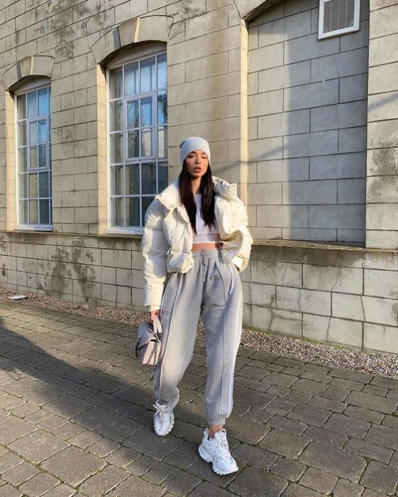 chica de cabello castaño largo usando una gorra gris, camisa blanca, abrigo blanco, pantalones deportivos pants grises, tenis blancos y bolsa de mano gris