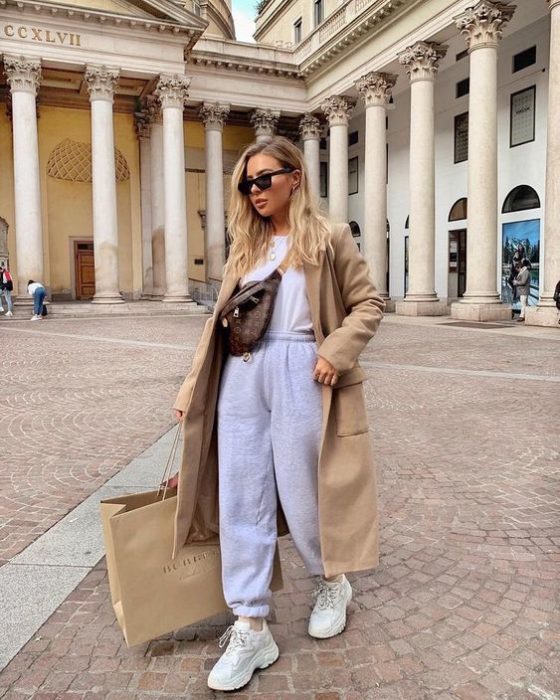 chica rubia con lentes de sol, de shopping, usando una bolsa tipo crossover de piel, camiseta blanca, abrigo beige, pants grises y tenis blancos deportivos