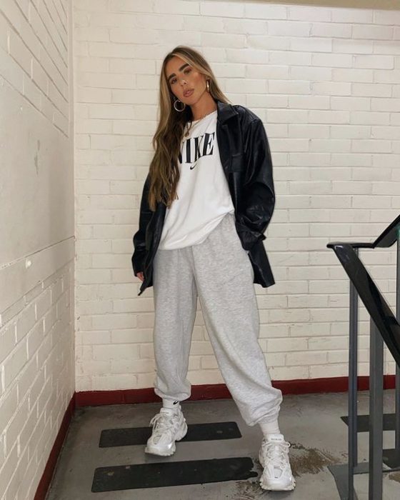 chica de cabello teñido rubio usa chaqueta larga de piel negro, camiseta blanca, pants grises y tenis blancos deportivos