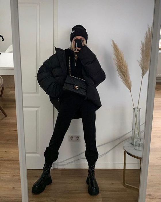 chica rubia tomandose un selfie con un beanie negro, chamarra oversized negra, top negro, bolso de piel negro, pantalones pants negros y botas de cuero