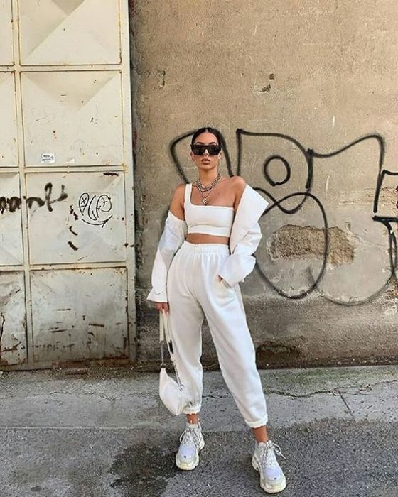 chica de cabello castaño usando lentes de sol, un top blanco de tirantes, blazer beige, pantalones pants blancos y tenis deportivos blancos