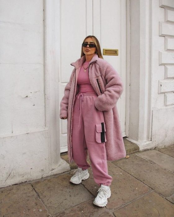 chica rubia usando lentes de sol, pants color rosa, un top rosa y abrigo de piel color rosa, tenis deportivos blancos