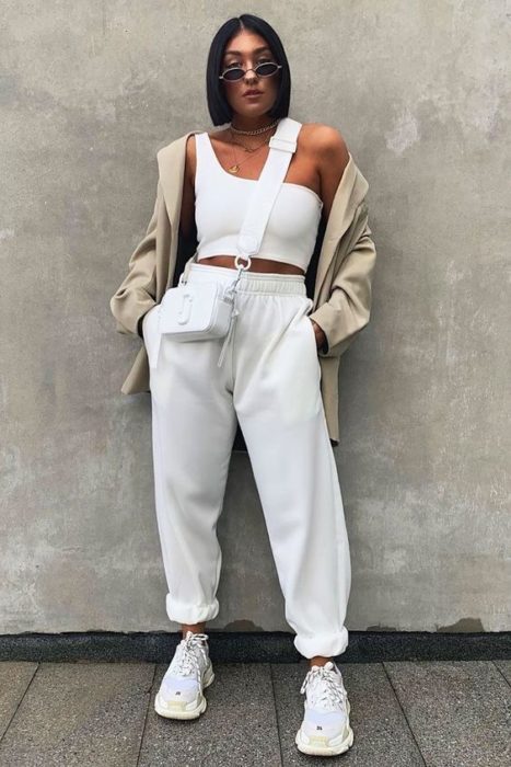 chica morena con cabello oscuro corto, usando lentes de sol, top de tirantes blanco, blazer beige, bolsa blanca de piel, pants blancos y tenis deportivos blancos