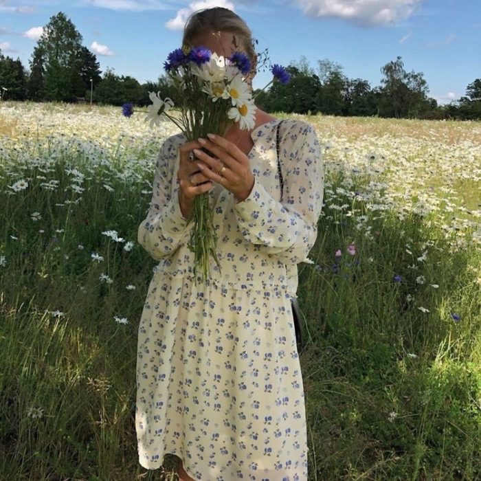 chica rubia posando con flores enfrente de la cara, usando un vestido amarillo pastel con estampado de flores en un campo abierto