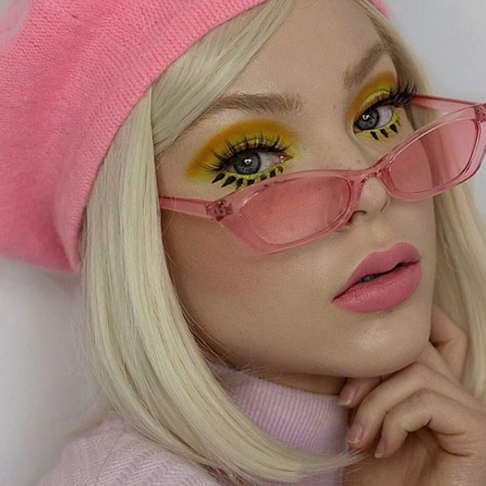 chica rubia usando una boina rosa, lentes de sol rosas, maquillaje amarillo con verde, delineado cat-eye negro y labial rosa