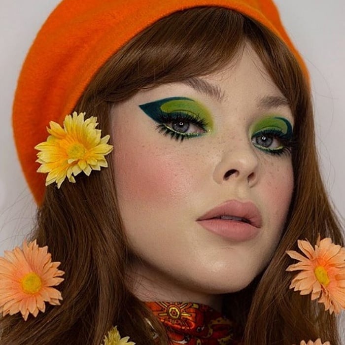 chica con cabello castaño largo usando maquillaje de sombra de ojos verde, delineado oscuro con flores naranjas en el cabello