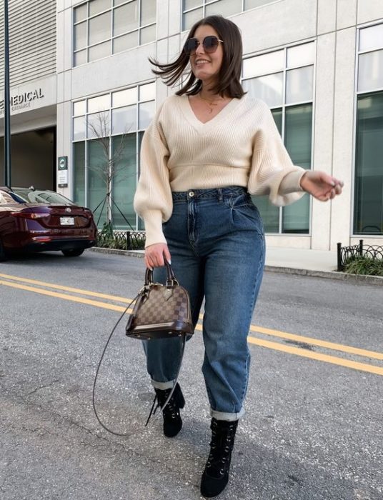 chica de cabello castaño corto usando lentes de sol, con jeans slouchy de mezclilla, suéter beige, botas negras y bolso de piel