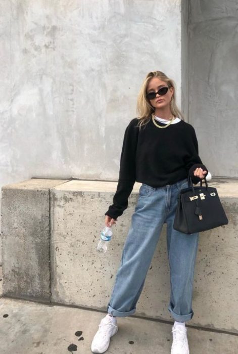 chica rubia con sudadera negra oversized, lentes de sol, bolso negro de piel y botella de agua