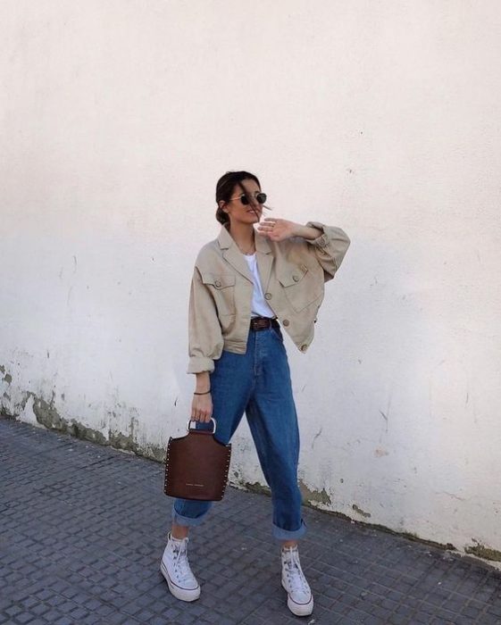 chica de cabello castaño usando lentes de sol, cazadora beige, playera blanca, bolso cafe de piel, jeans slouchy y tenis deportivos blancos