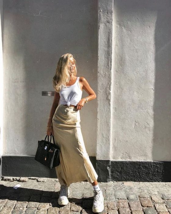 chica rubia con top de tirantes blanco, falda midi de satén amarillo pastel y tenis deportivos, bolso negro de piel