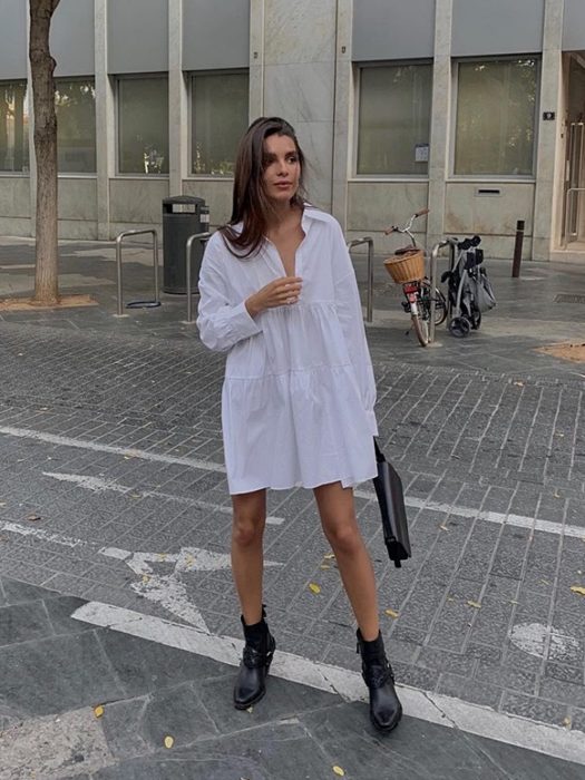 chica con vestido blanco largo, o camisa de vestir oversized, botas negras y cabello castaño, bolso de mano negro