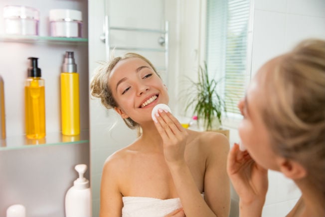 Chica frente al espejo sonriendo y limpiando su rostro con una almohadilla de algodón