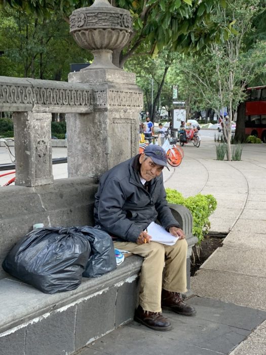 Don Baldemar, abuelito sentado en una banca de cemento dibujando en una libreta blanca, vende dibujos para sobrevivir