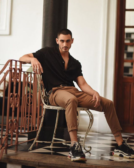 Alejandro Speitzer posando para fotografía, sentado en una silla, usando jogger cakis y camisa negra con tennis