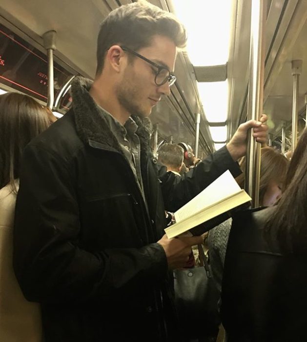 Hombre con gafas sostenido de una barra de metal y leyendo un libro