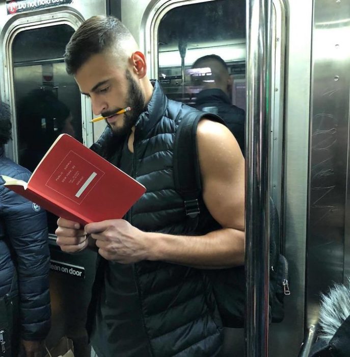 Hombre llevando chaleco deportivo leyendo un libro rojo y sosteniendo un lápiz entre sus labios 