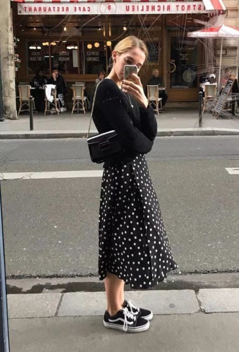 Chica rubia toándose una selfie con suéter y falda negros con tenis negros vans