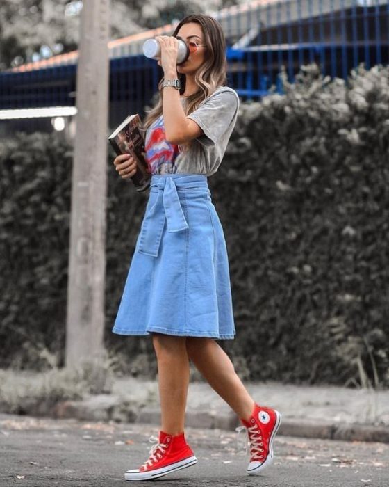 Chica rubia bebiendo café con falda midi de mezclilla, converse rojos y blusa gris