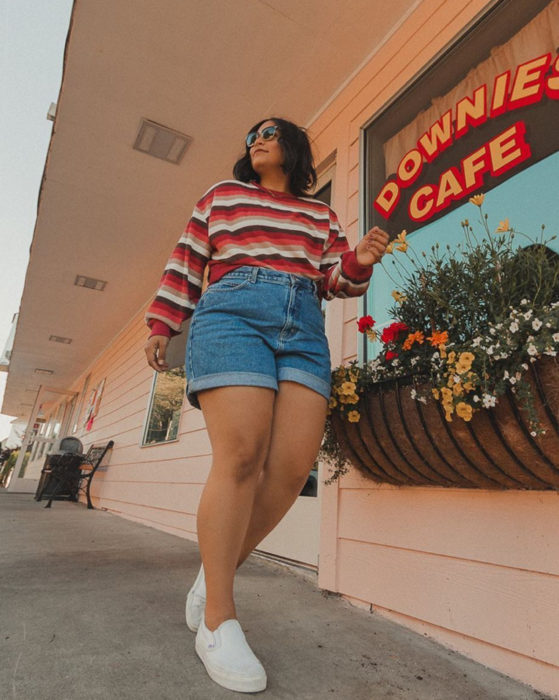 Looks con short vintage; mujer plus size caminando en la calle y sonriendo, cabello negro y corto, lentes de sol, sudadera de rayas rojas, blancas y café, tenis blancos