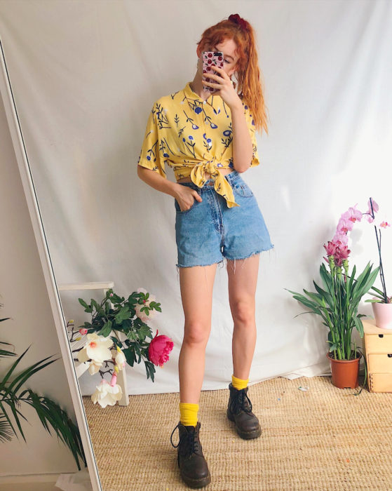 Looks con short vintage;chica pelirroja tomándose selfie frente al espejo, con blusa de flores amarilla y botas Dr. Martens