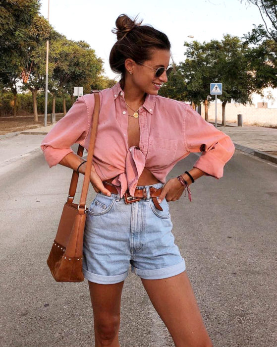 Looks con short vintage; mujer sonriendo en la calle, con blusa de mezclilla rojo deslavado o rosa salmón y bolsa color café, peinado de chongo alto y lentes de sol, manos en la cintura