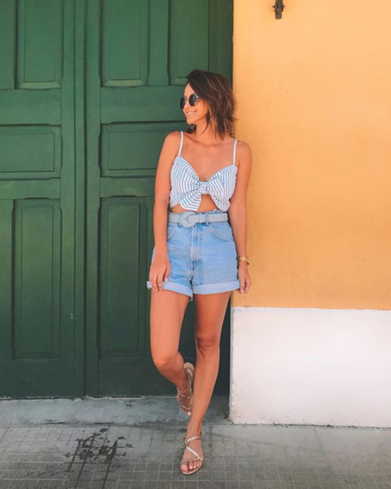 Looks con short vintage; mujer recargada en la pared frente a una puerta antigua verde, cabello corto a los hombros, estilo playero, top rayado con moño, sandalias doradas