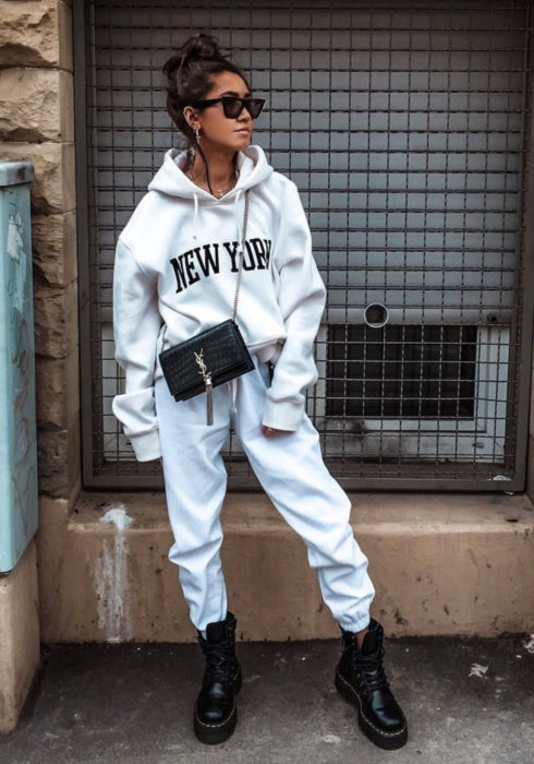 Looks con sudadera; mujer en la calle con peinado de chongo alto, lentes de sol modernos, hoodie blanco con estampado de Nueva York, pants, bolsa negra, botas militares Dr. Martens