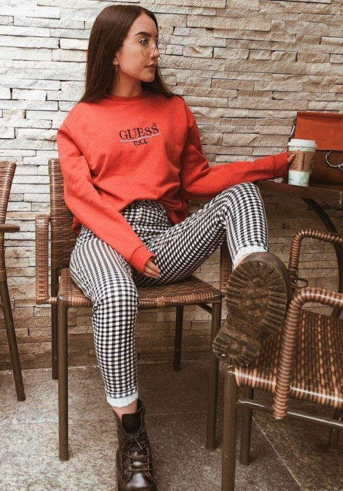 Looks con sudadera; mujer sentada en silla de restaurante, con cabello largo, lacio y castaño rojizo, hoodie rojo, pantalón a cuadros blanco y negro y botas Dr. Martens