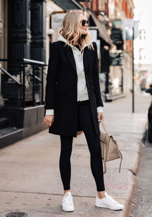 Looks con sudadera; mujer en la calle, cabello rubio, corto y ondulado, lentes de sol oscuros, hoodie gris, abrigo mediano negro, leggins, tenis blancos y bolso beige