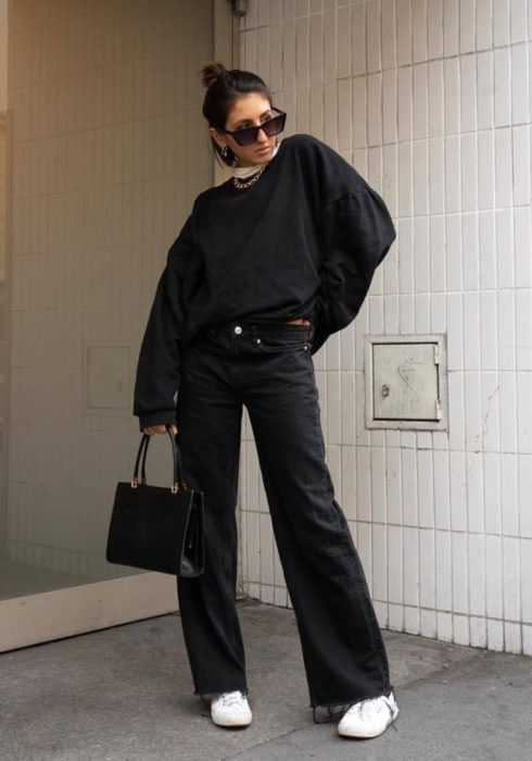 Looks con sudadera; mujer de cabello corto castaño con peinado de media cola, lentes de sol grandes, hoodie negro oversized, pantalones jeans acampanados, tenis blancos y bolsa de mano