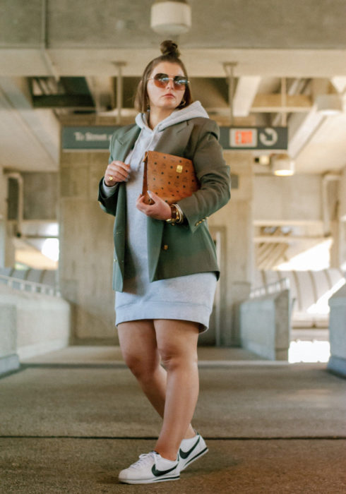 Looks con sudadera; chica plus size usando hoddie gris como vestido, con saco verde militar, tenis Nike blancos y bolsa de cartera café; peinado para cabello corto peinado con medio chongo alto