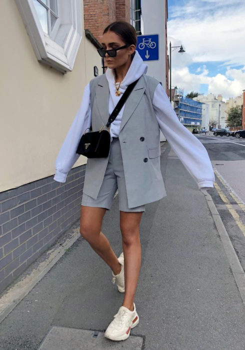 Looks con sudadera; mujer caminando en la calle con lentes de sol, vestida con hoodie azul cielo, chaleco y short color gris, tenis deportivos beiges, bolsa negra